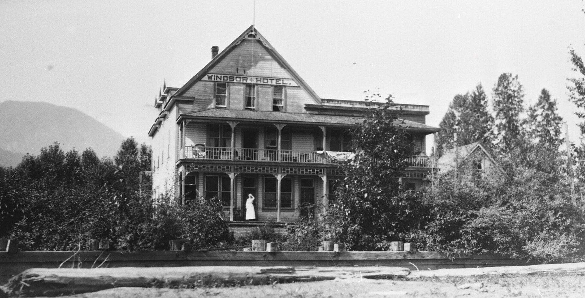 In a photograph taken from the banks of Lardeau Creek in the early 20th century, the Windsor Hotel is visible, along with a woman standing near its front door. The woman is likely the hotel’s proprietor Alice Jowett. Photo: Courtesy of Arrow Lakes Historical Society