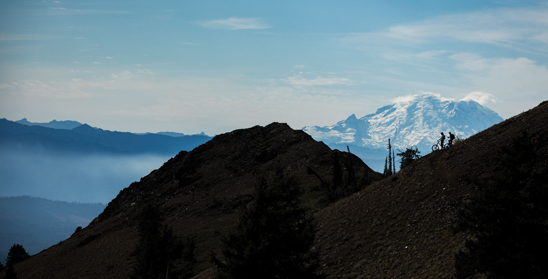 Mount Rainier dwarfs Aric Duncan and Michael Fischer as they take in the view before ripping down a golden hillside meadow.
